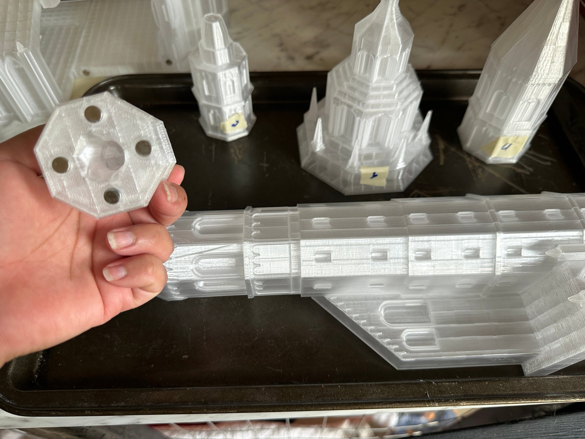 Hand holding a clear plastic component with other similar components on a black tray.