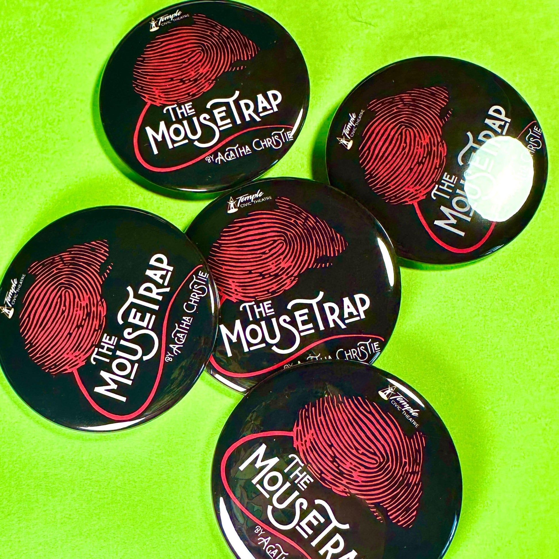 Five 'The Mousetrap' buttons by Agatha Christie on a green background