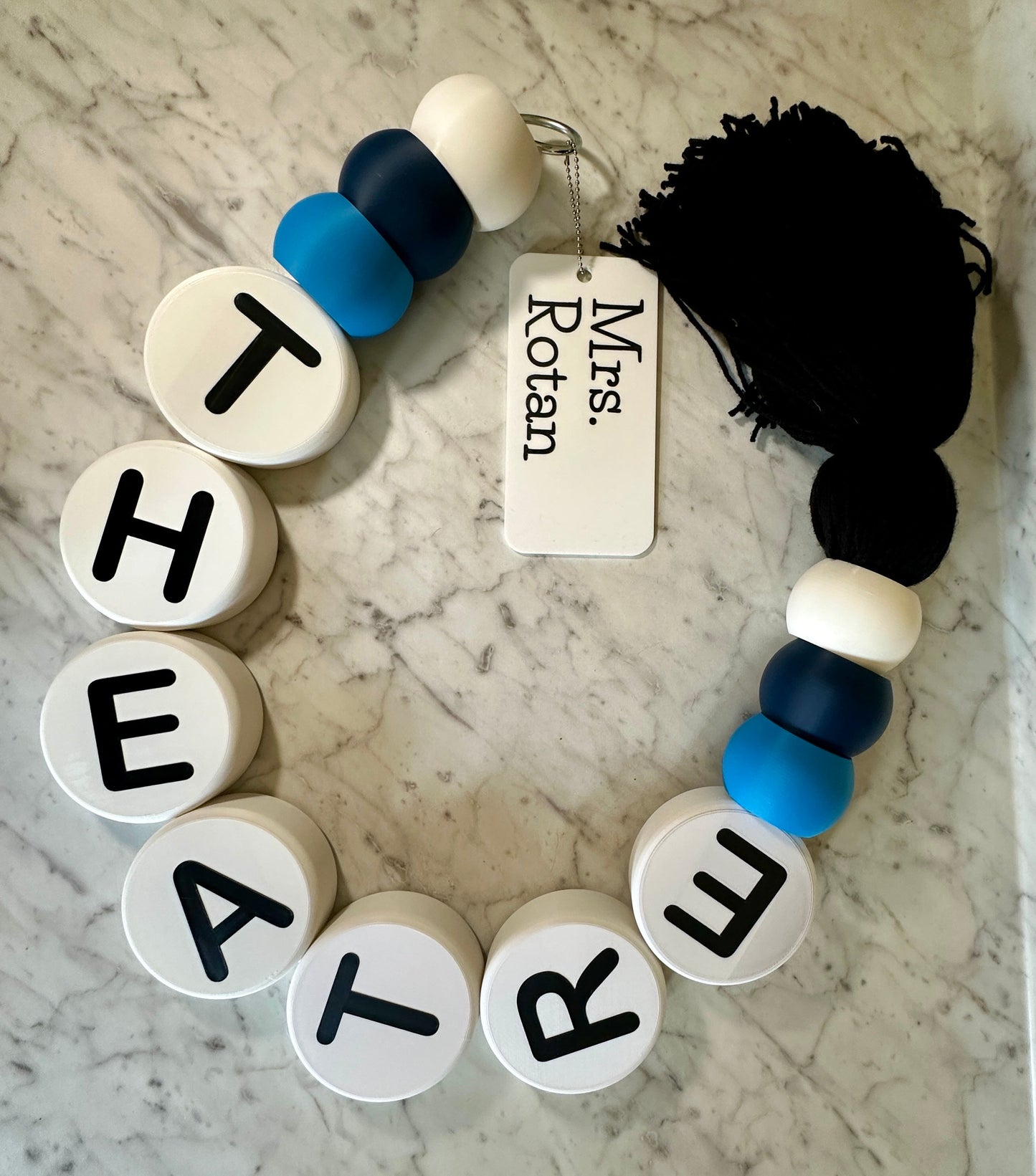 Bracelet with letter beads spelling 'THEATRE' on a marble surface with a tag labeled 'Mrs. Rotan'.