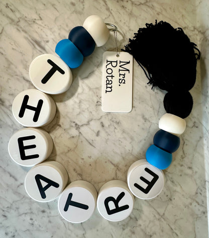 Bracelet with letter beads spelling 'THEATRE' on a marble surface with a tag labeled 'Mrs. Rotan'.