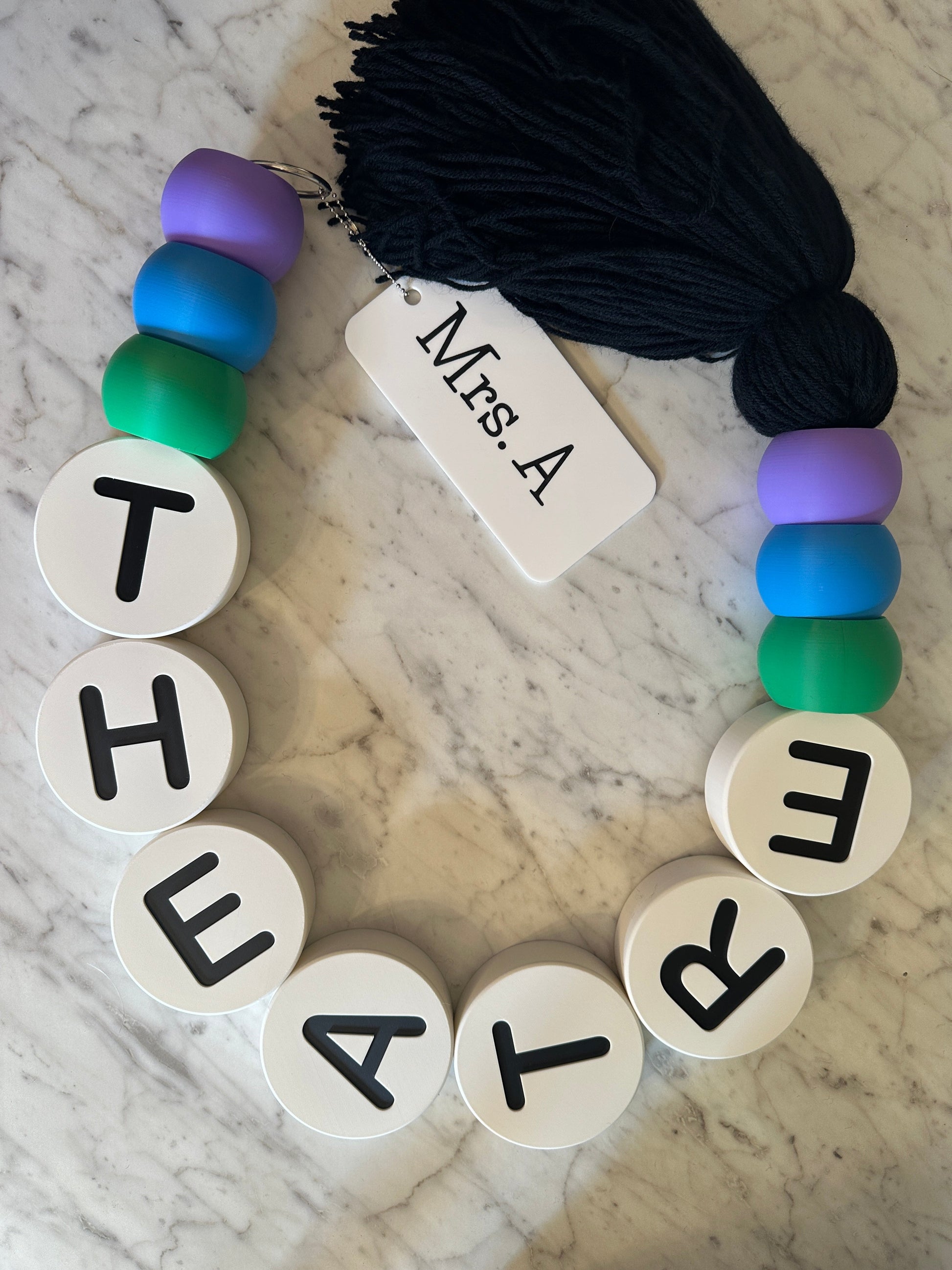 Bracelet with letter beads spelling 'THEATRE' on a marble surface with yarn.