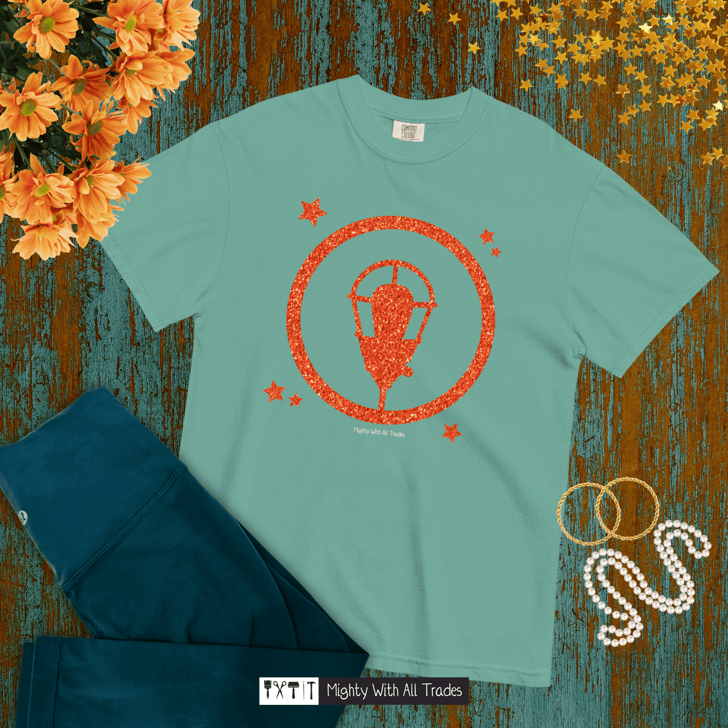 Teal t-shirt with orange glitter ghost light graphic design on a textured surface with flowers and jewelry.