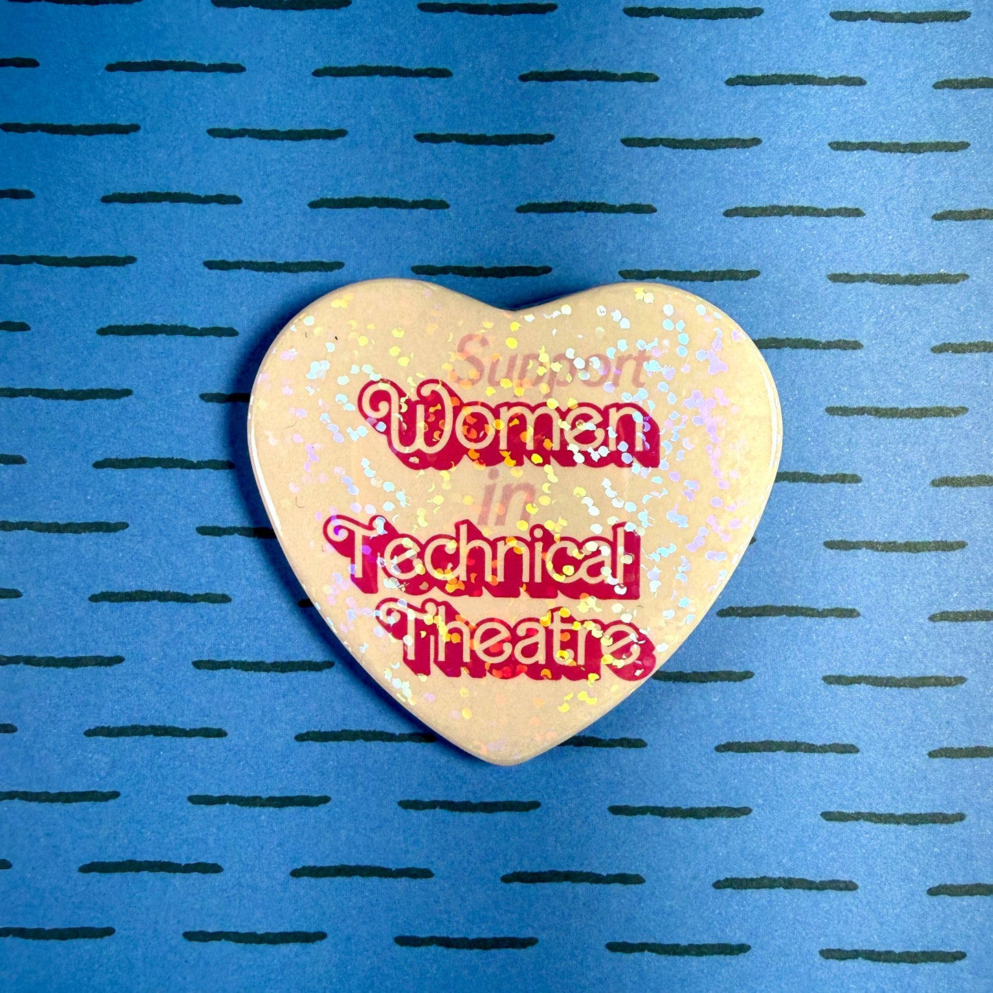 Heart-shaped pin with 'Support Women in Technical Theatre' text on a blue background